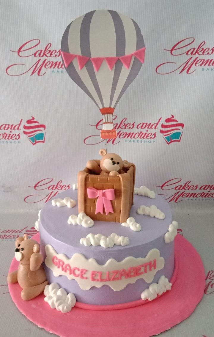 Purple hot air balloon cake with fondant bears and white icing clouds on a pink base board.