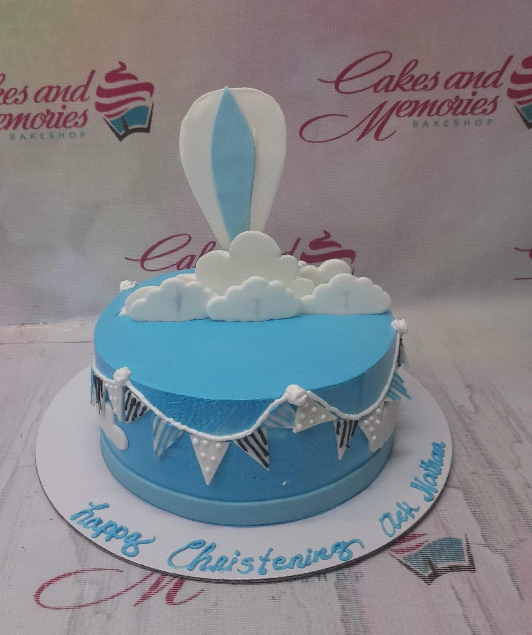 Light blue christening cake with a hot air balloon topper, white fondant clouds, and decorative bunting on the side.