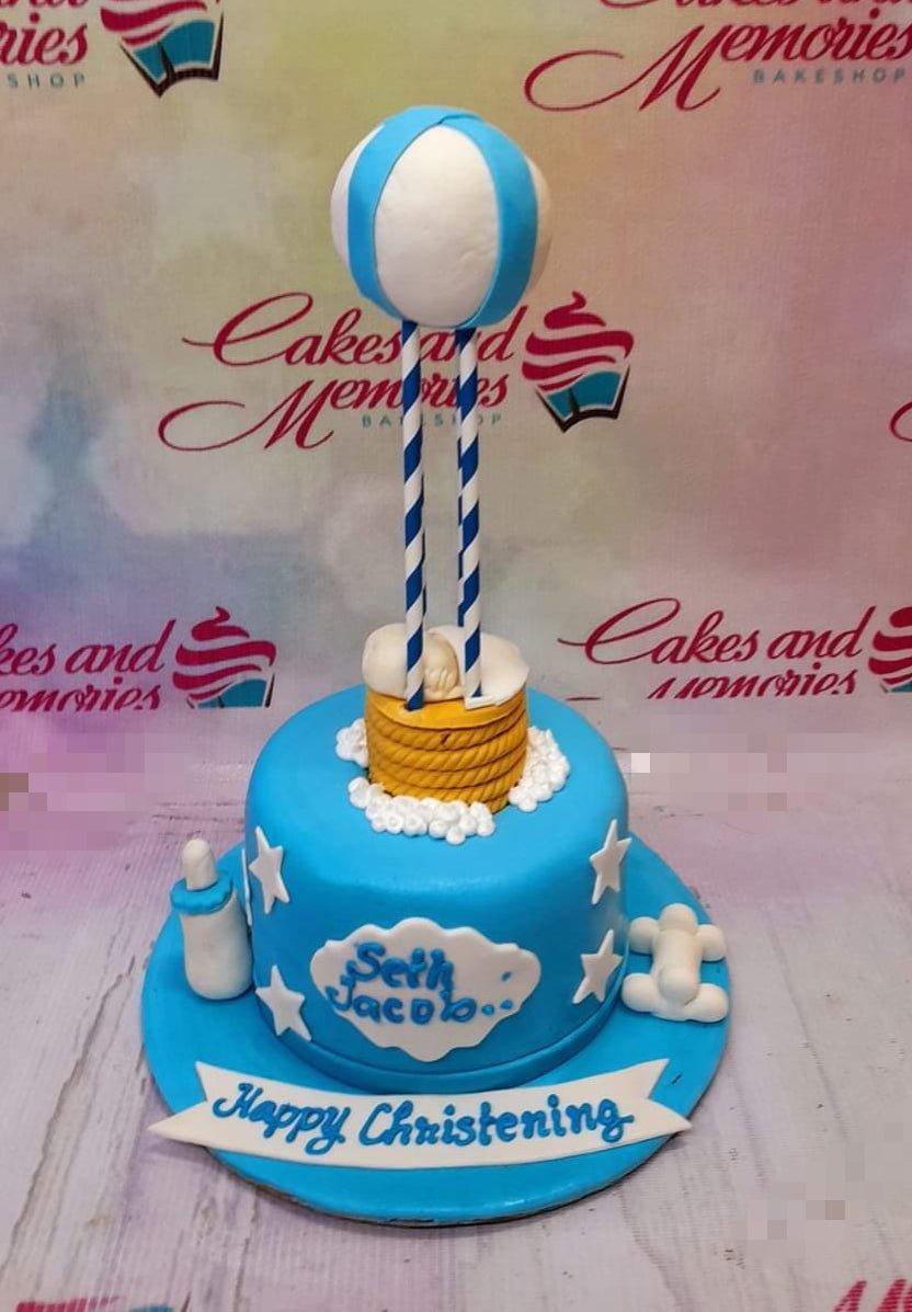 Blue hot air balloon christening cake with a sleeping baby figurine, white clouds, and stars on a blue fondant base.
