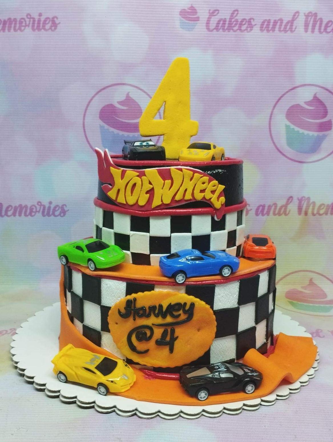 Two-tier Hot Wheels themed birthday cake with checkered racing flag pattern, toy cars, and a yellow number four topper.