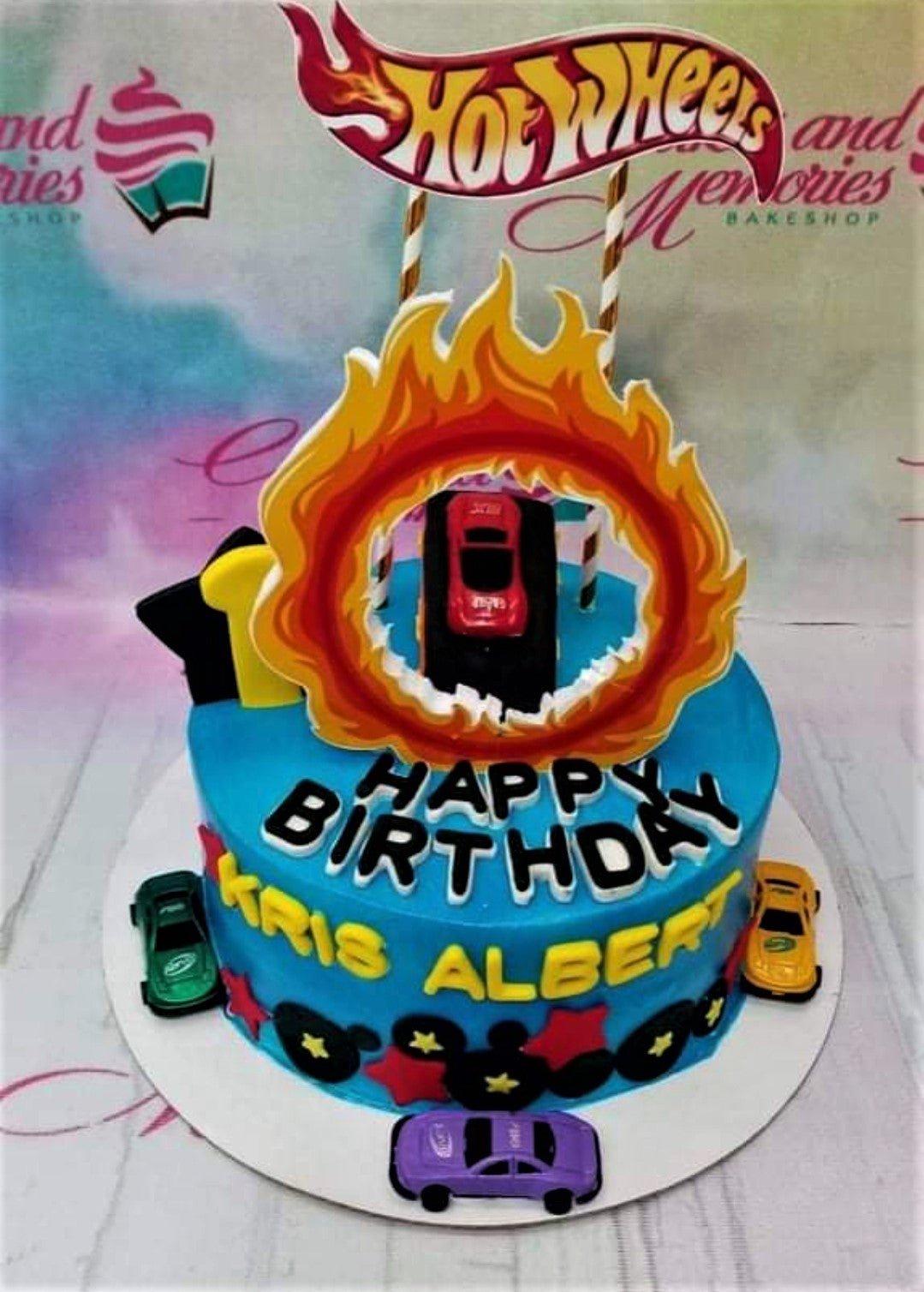 Blue Hot Wheels themed birthday cake with toy cars, a ring of fire topper, and black gumpaste letters for Kris Albert.