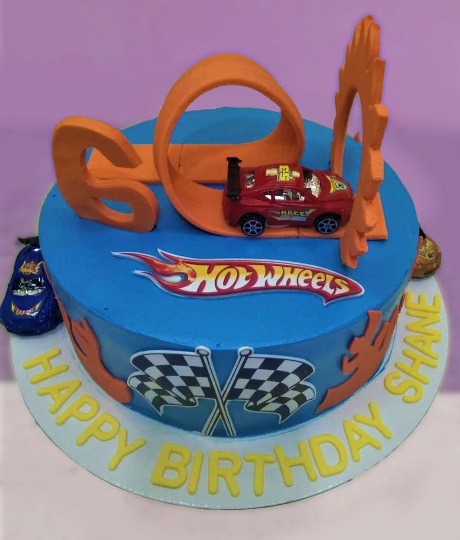Blue Hot Wheels themed birthday cake with orange racing tracks, toy cars, and checkered flags.