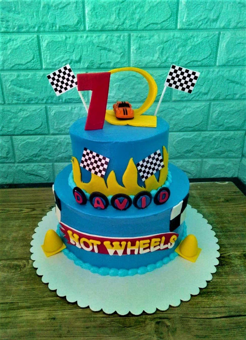 Two-tier blue Hot Wheels themed birthday cake with racing flags, yellow flames, and a toy car topper.