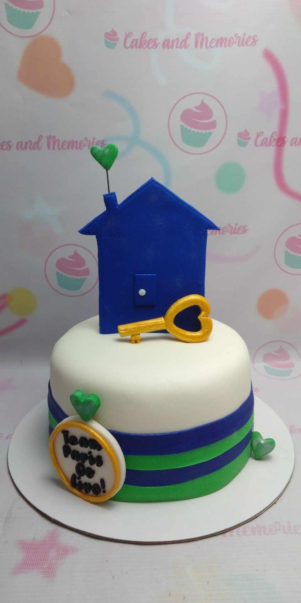 White fondant cake with a large blue house topper, a gold heart key, and green heart accents with a custom message plaque.