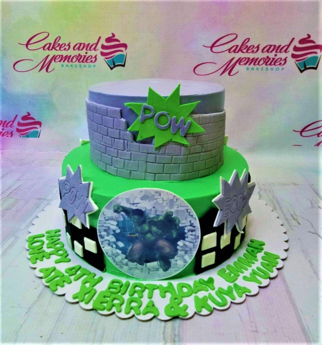 Two-tier Hulk themed birthday cake with green and purple fondant, brick wall design, and Hulk printout topper.