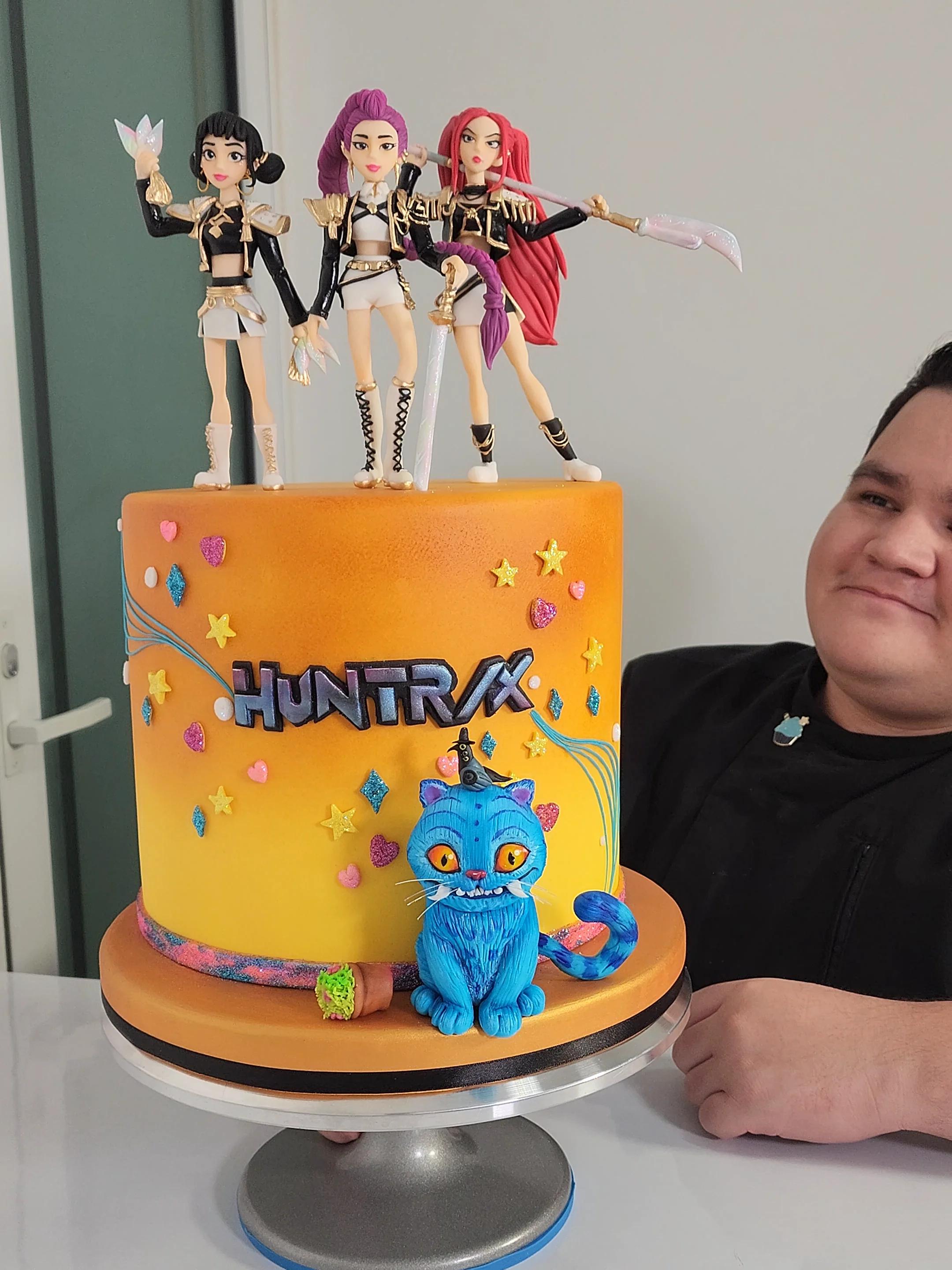 Tall orange and yellow gradient fondant cake featuring three anime-style warrior women figurines and a blue cat with a small bird on its head.