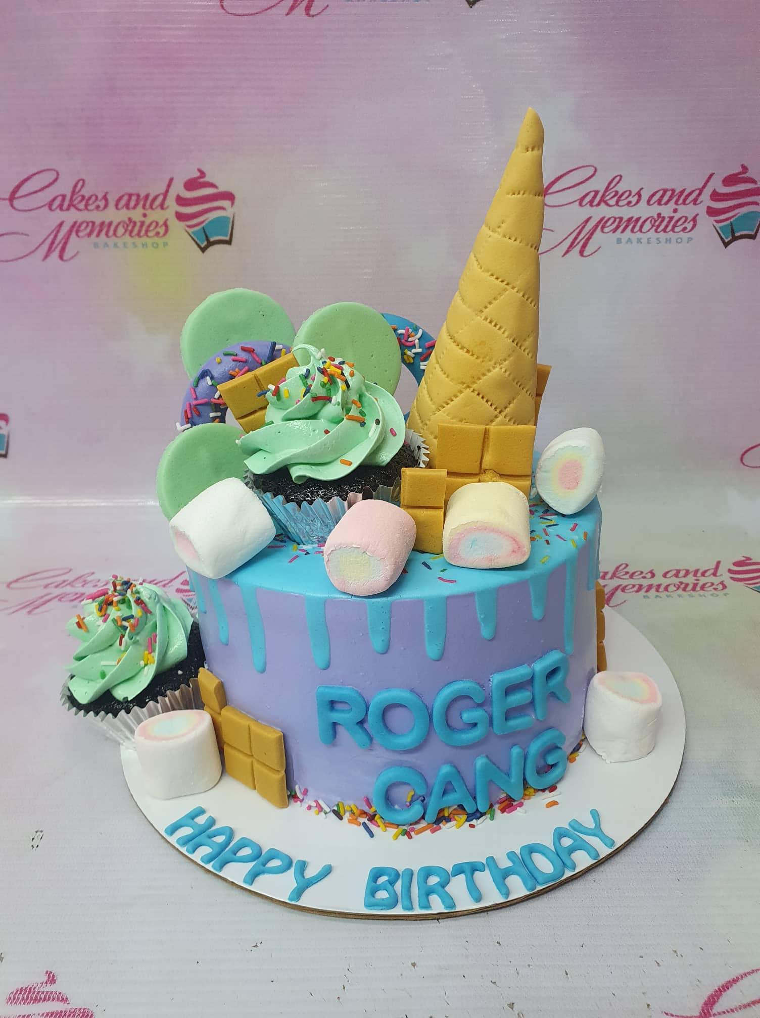 Purple and blue birthday cake with an ice cream cone topper, chocolate bars, marshmallows, and cupcakes.