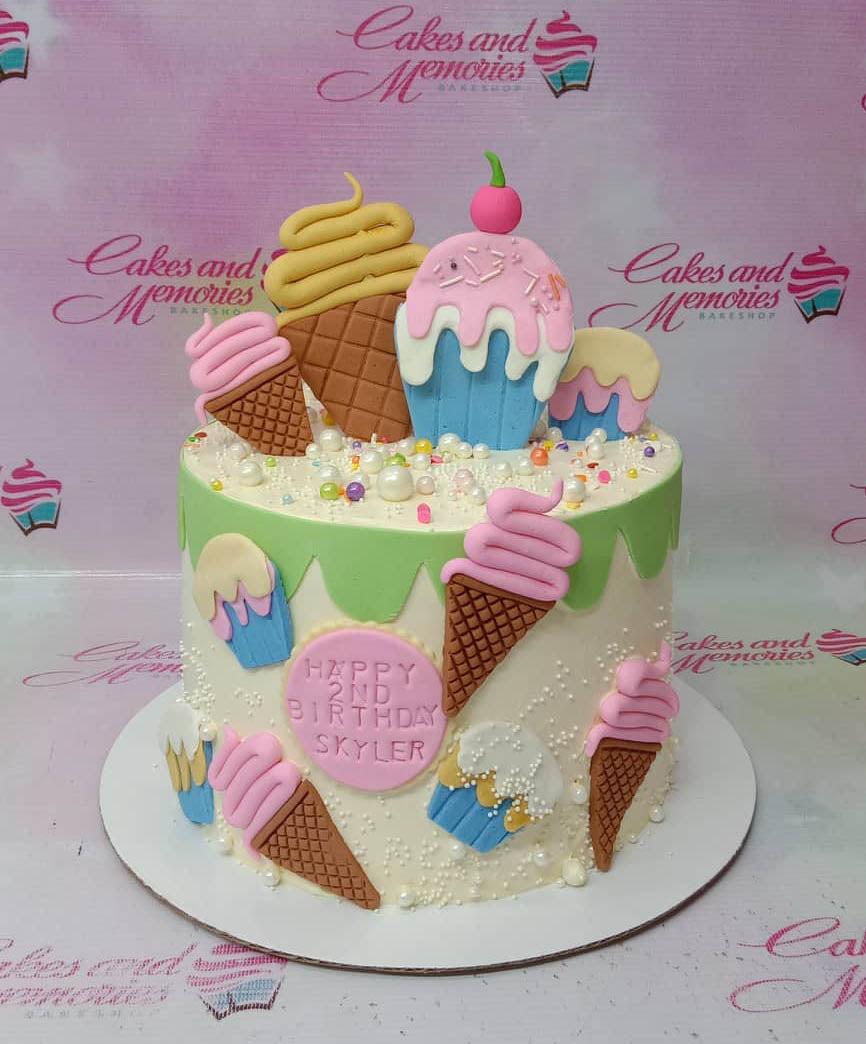 White 1-tier birthday cake with green icing drip, fondant ice cream cones, cupcakes, and colorful sprinkles.
