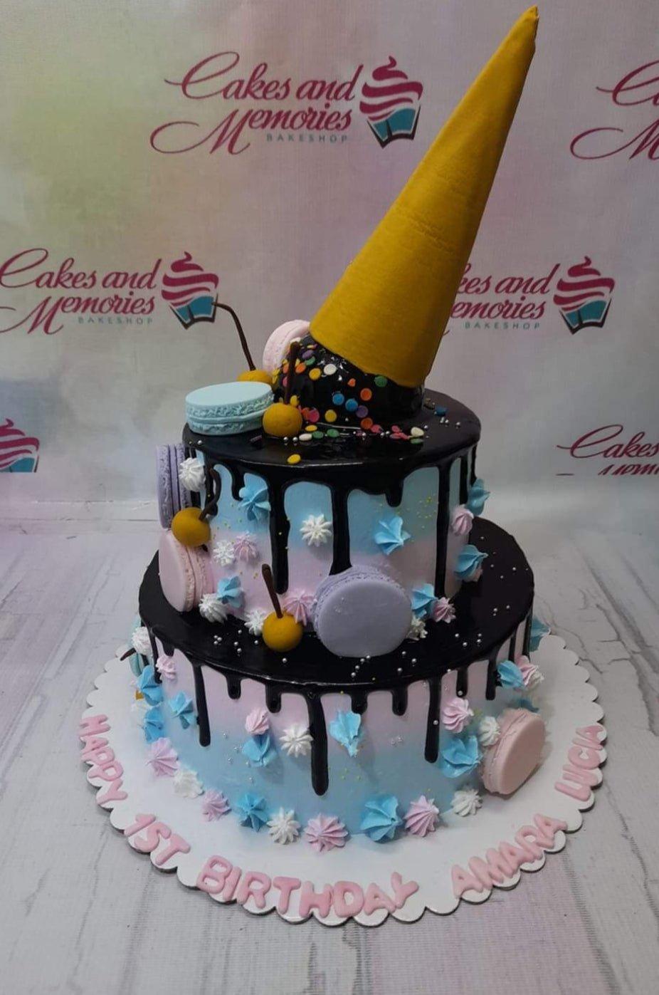 Two-tier pastel blue and pink gradient cake with chocolate drip, ice cream cone topper, macarons, and colorful icing rosettes.