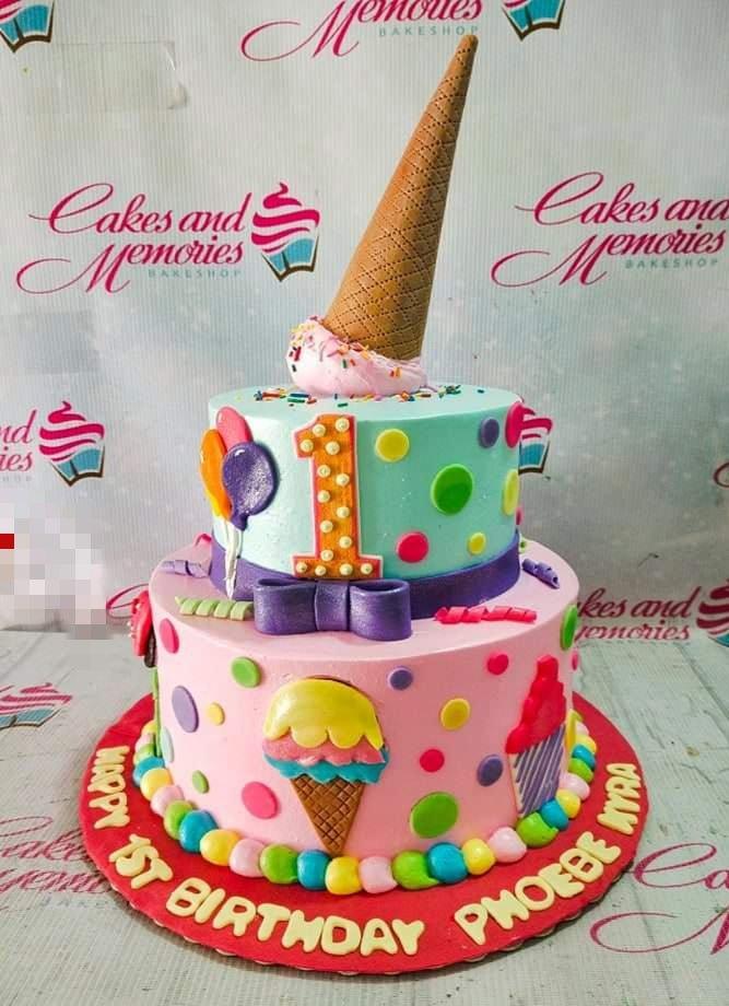 Two-tier pink and light blue ice cream themed birthday cake with a large waffle cone topper and colorful fondant decorations.