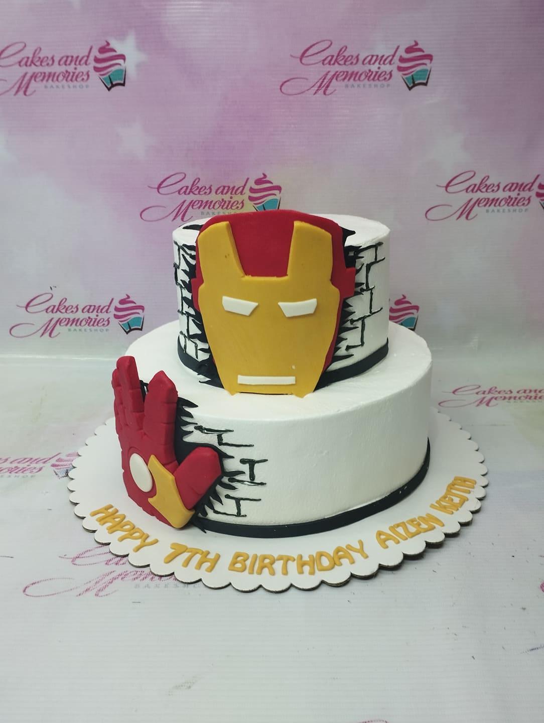 Iron Man themed 2-tier white cake with red and yellow fondant mask and hand toppers and black brick wall icing details.