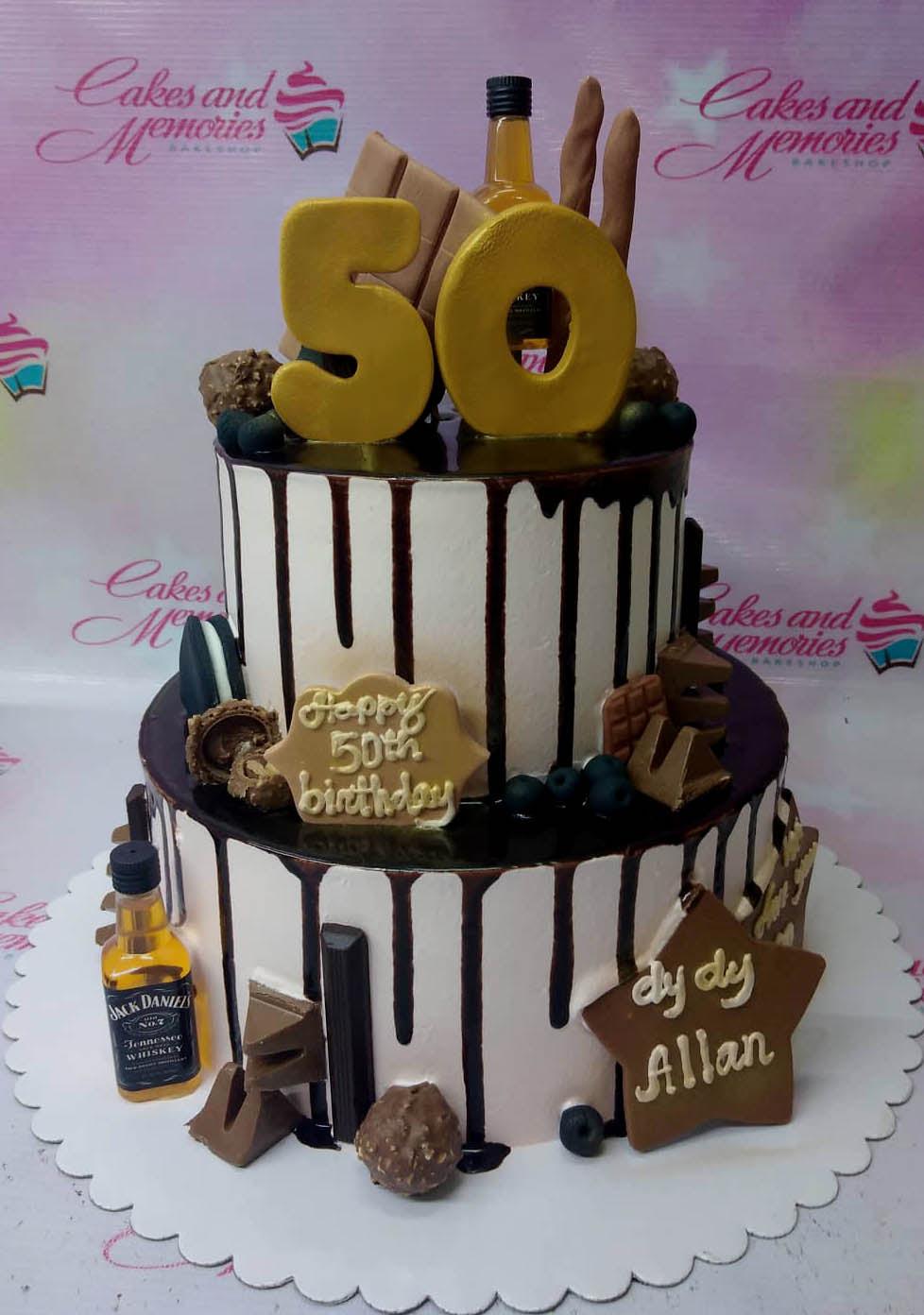 Two-tier white cake with chocolate drip, Jack Daniels bottle, chocolate bars, and gold 50 topper for a 50th birthday celebration.