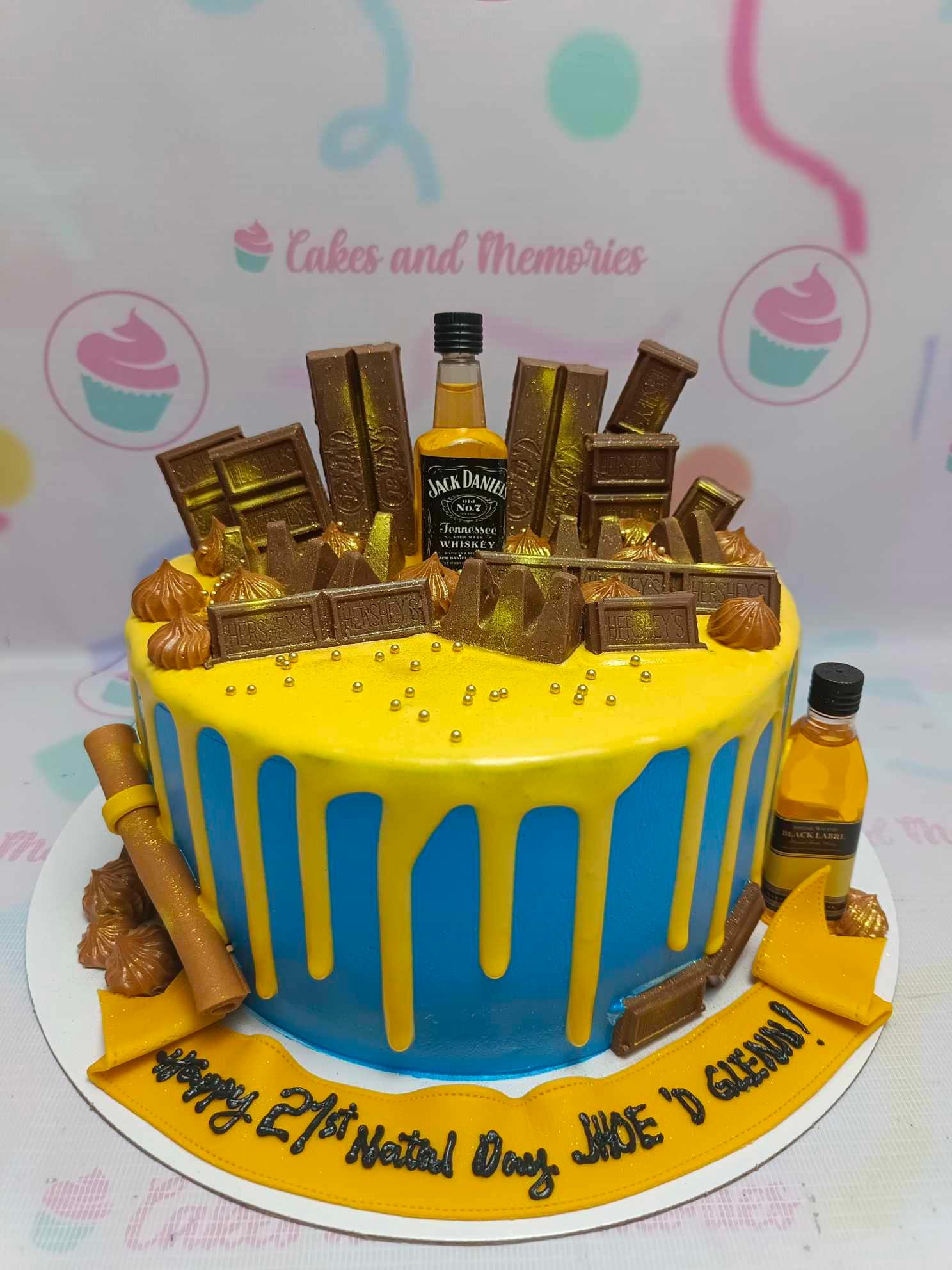 Blue and yellow drip cake with Jack Daniel's and Black Label mini bottles, Hershey's and KitKat chocolates, and gold dragees.
