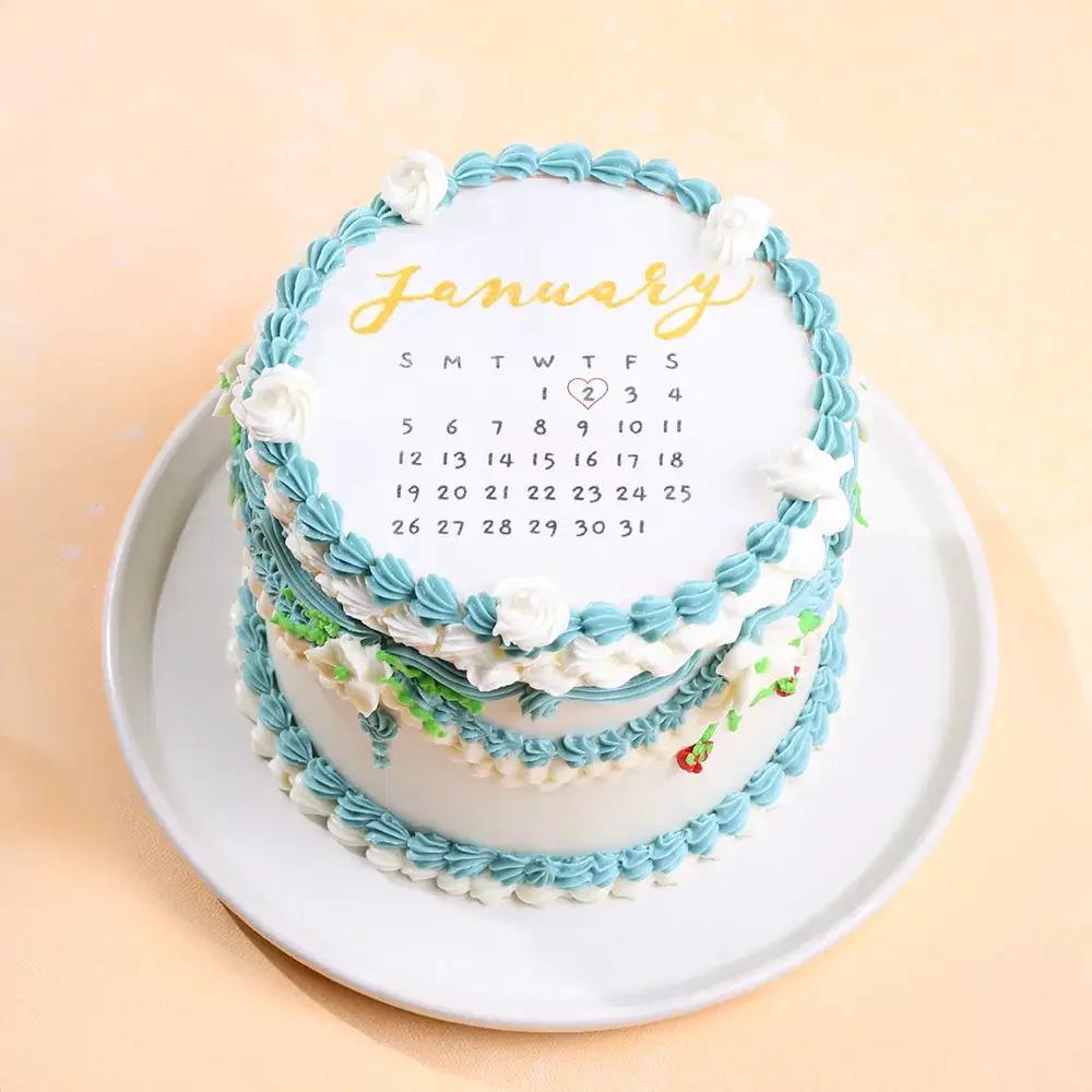 White minimalist bento-style calendar cake with blue and white piped icing borders, featuring a January calendar design and small white roses.