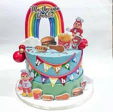 Two-tier Jollibee themed birthday cake with blue and green icing, rainbow topper, and various fast food printouts.