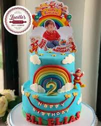 Two-tier blue Jollibee themed birthday cake with rainbow, clouds, and character toppers for Eli Elias's 2nd birthday.