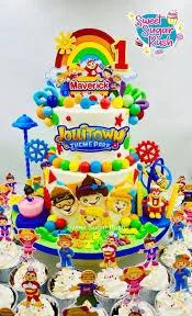 Colorful 2-tier Jollitown theme park birthday cake with rainbow, sun, and character printouts on a blue base board.