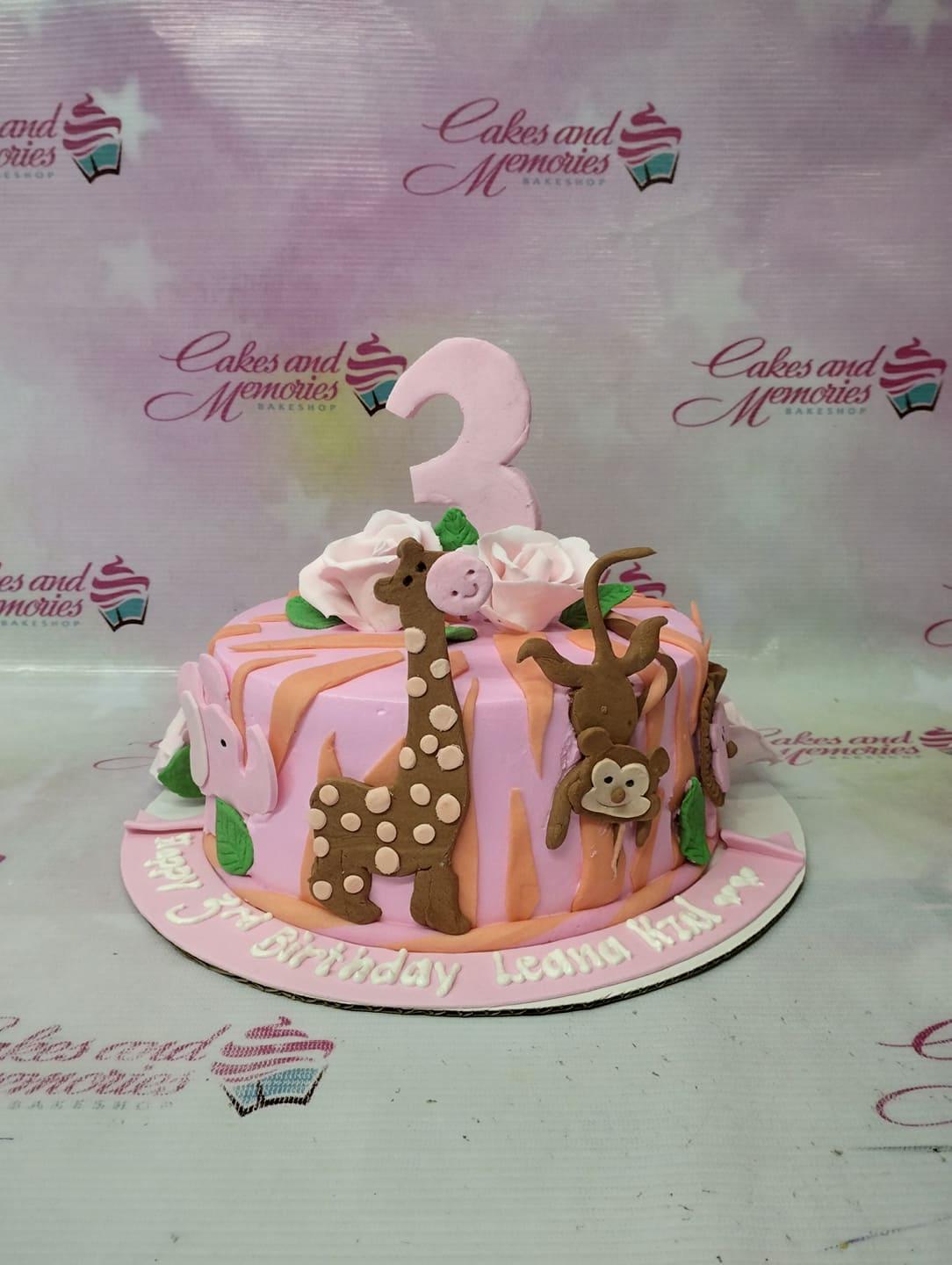 Pink jungle themed birthday cake with fondant giraffe, monkey, and elephant figures, topped with a large pink number 3 and roses.