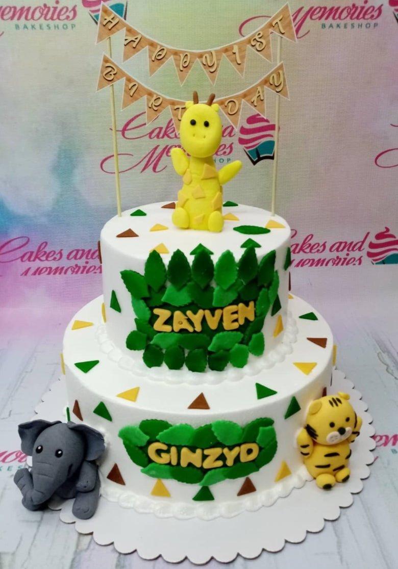 White 2-tier jungle animal birthday cake with fondant giraffe, elephant, and tiger figures and green leaf decorations.