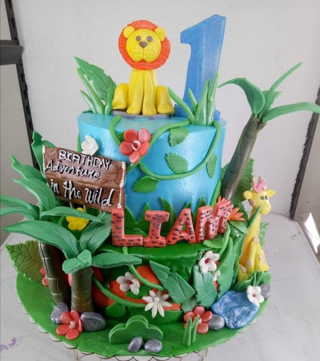 Two-tier jungle safari birthday cake with fondant lion, giraffe, palm trees, and 'Liam' name blocks.