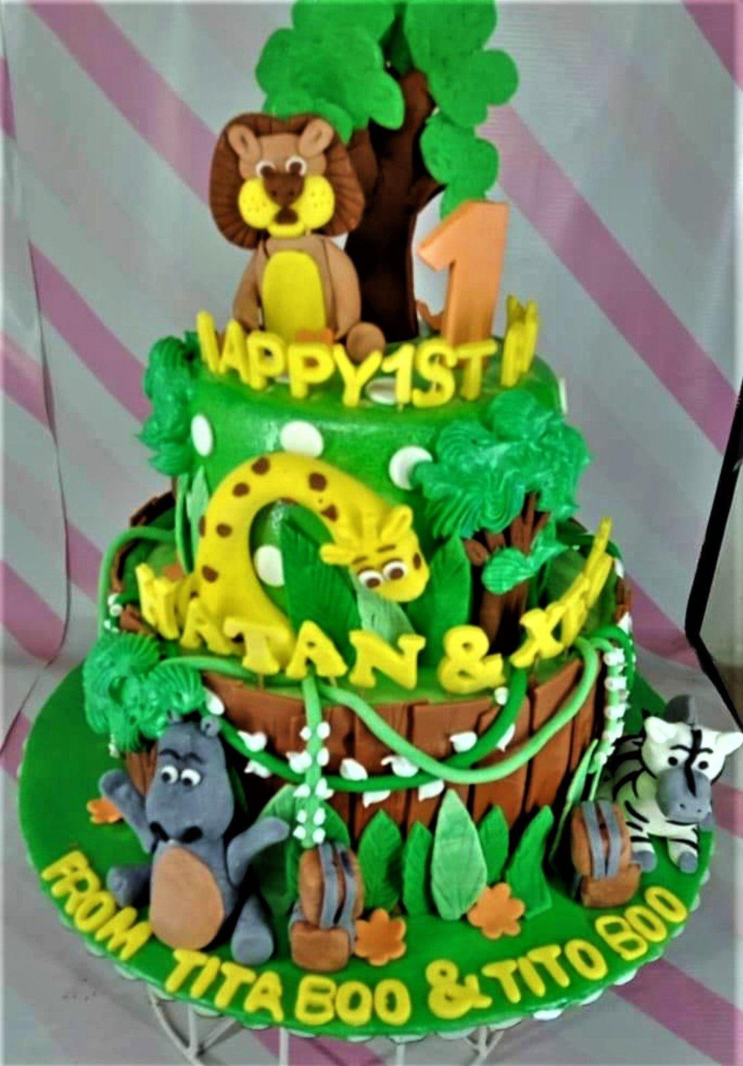 Three-tier jungle safari themed fondant cake with 3D edible lion, giraffe, hippo, and zebra figures, green foliage, and yellow lettering.