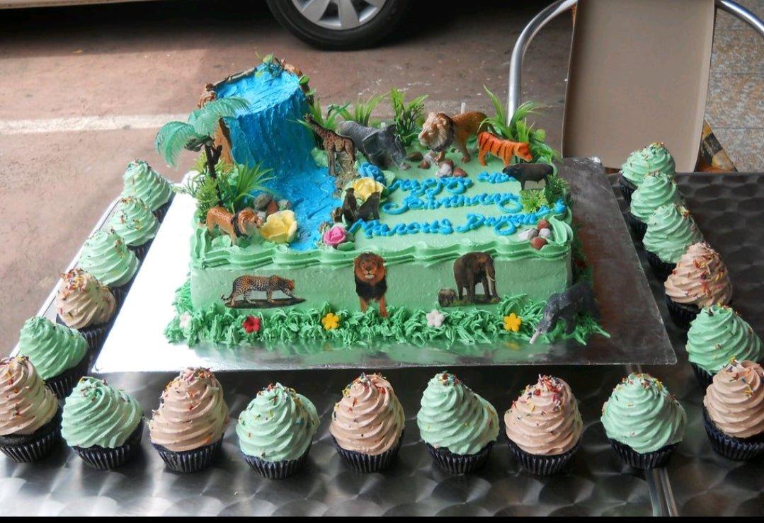 Jungle safari themed rectangle cake with a blue icing waterfall, plastic animal toys, and green grass icing borders surrounded by cupcakes.