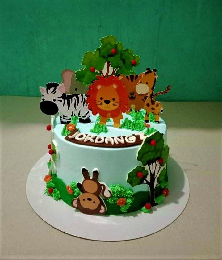 Light blue jungle safari cake with printout animals including a lion, zebra, giraffe, and monkey with green icing grass and trees.