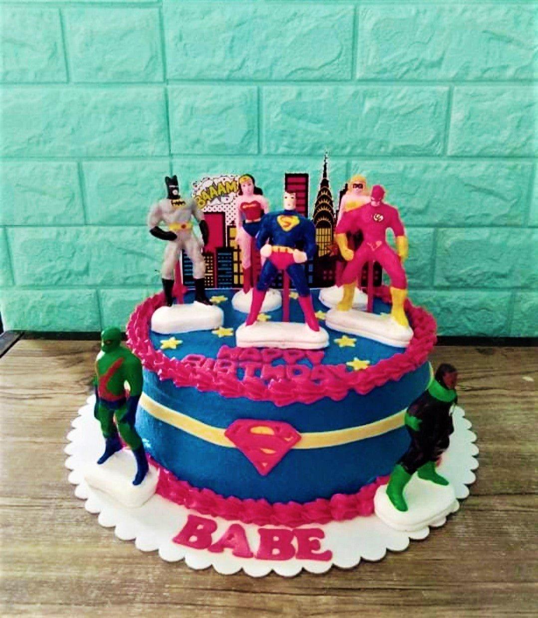 Blue Justice League superhero birthday cake with Batman, Superman, Wonder Woman, and Flash figurines and pink icing borders.