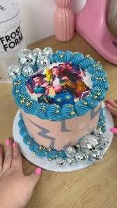 Pink and blue K-pop themed cake with a printed character topper, blue icing swirls, and silver disco ball decorations.