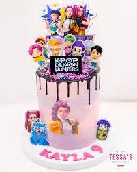 Pink K-Pop Demon Hunters themed cake with character printouts, black chocolate drip, and pink name message on the base.