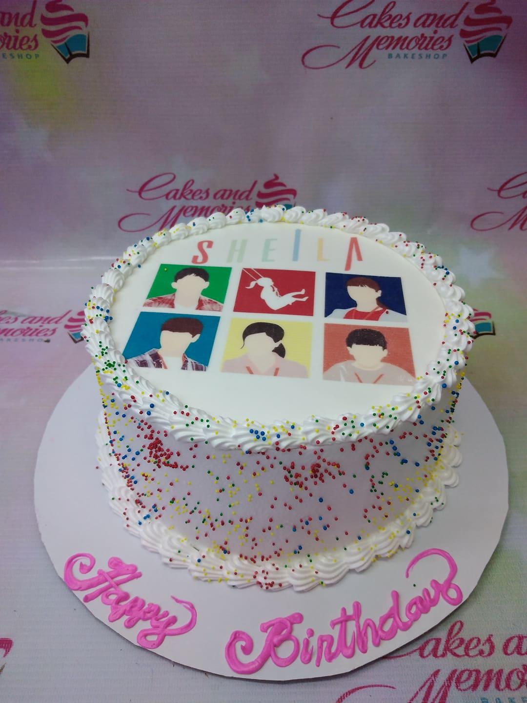 White round cake with a colorful K-pop group photo print on top, rainbow sprinkles on the sides, and pink icing text on the base board.