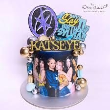 Black and blue Katseye K-pop themed cake with printed group photo, gold glitter toppers, and metallic gold and silver spheres.