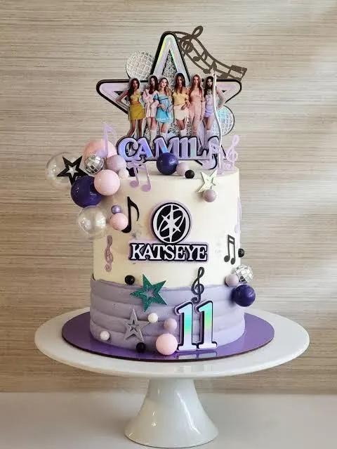 White and lavender tiered cake with Katseye K-pop theme featuring character printouts, musical notes, and stars.