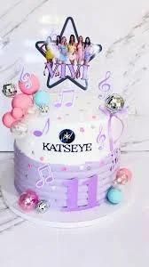 White and purple K-pop themed cake for Vivi's 11th birthday featuring Katseye group printouts and musical note decorations.
