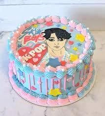 Light blue K-pop themed birthday cake with a hand-drawn style male character, pink and blue icing swirls, and colorful stars.