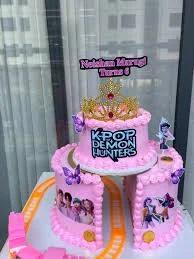 Pink two-tier K-pop and Demon Hunter themed birthday cake with a gold crown, purple butterflies, and character printouts.