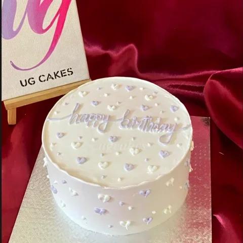 White minimalist birthday cake with lavender and white piped icing hearts and cursive Happy Birthday message.