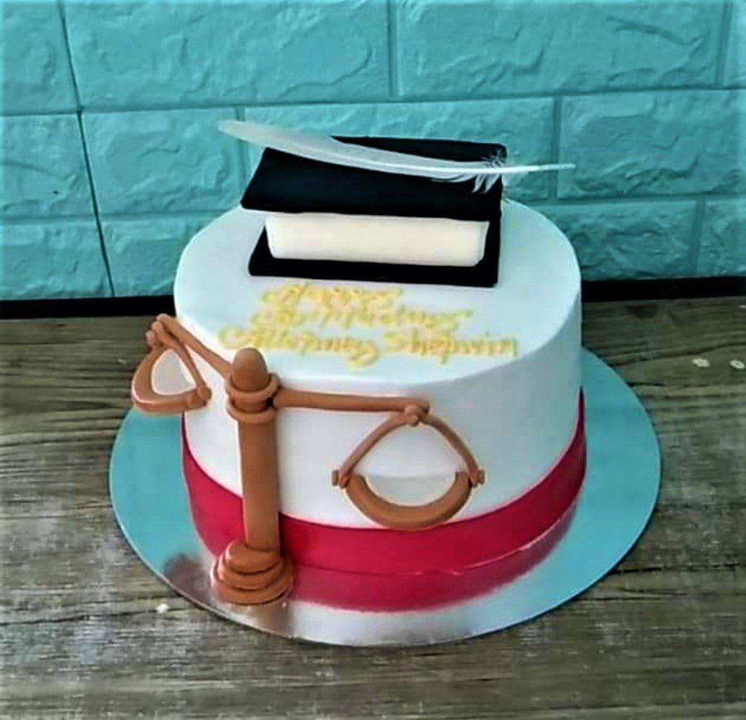 White fondant law graduation cake featuring a 3D book with a feather quill and a brown scales of justice topper.