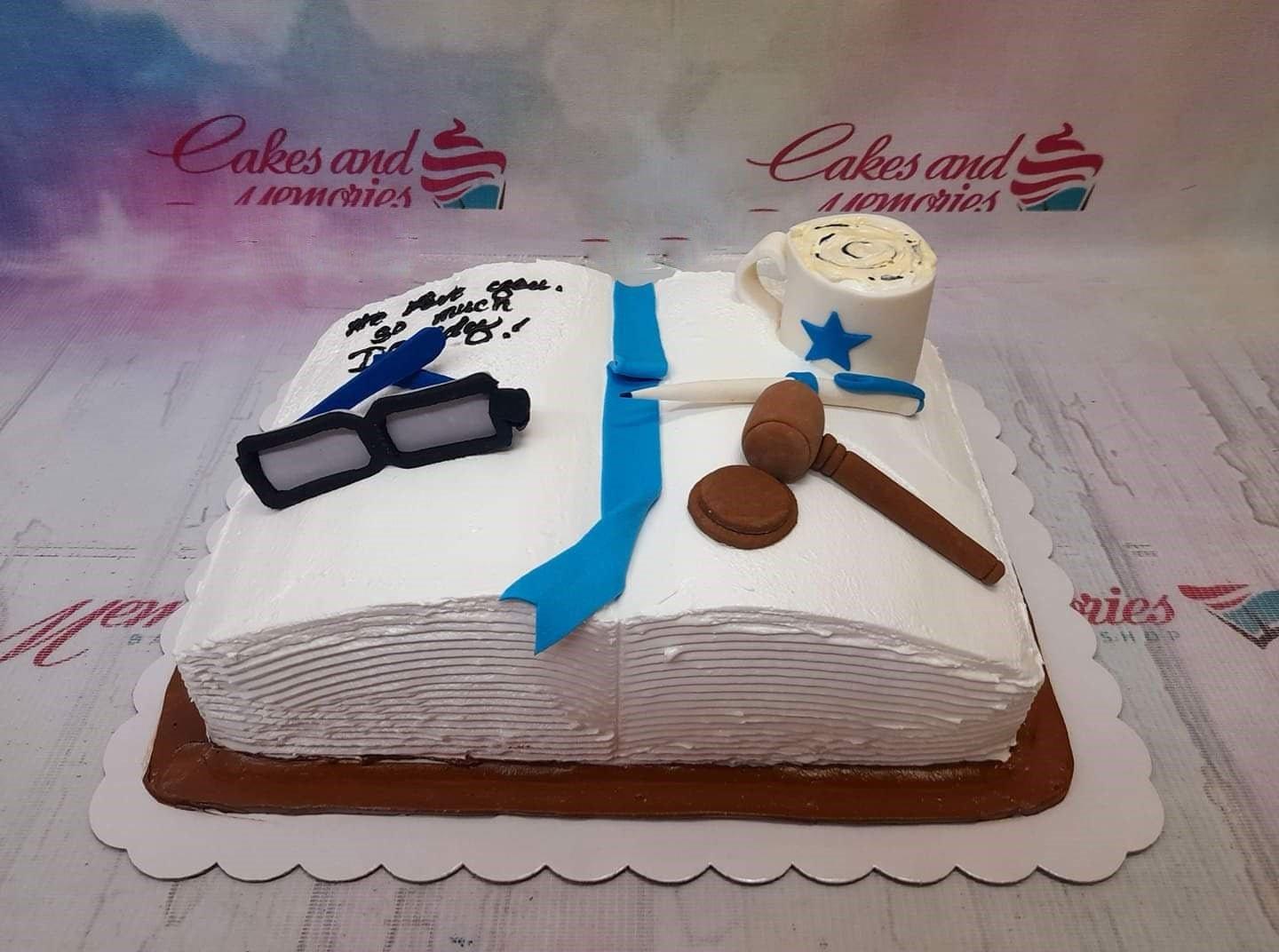 White rectangle book-shaped cake with edible 3D fondant glasses, a gavel, a pen, and a blue ribbon bookmark.