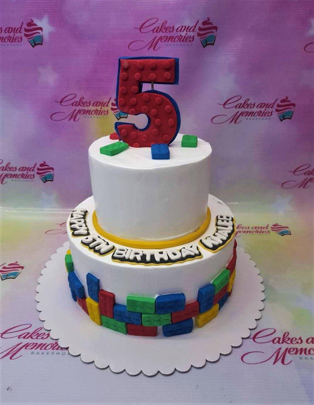 Two-tier white fondant Lego-themed birthday cake with colorful building block decorations and a large red number 5 topper.