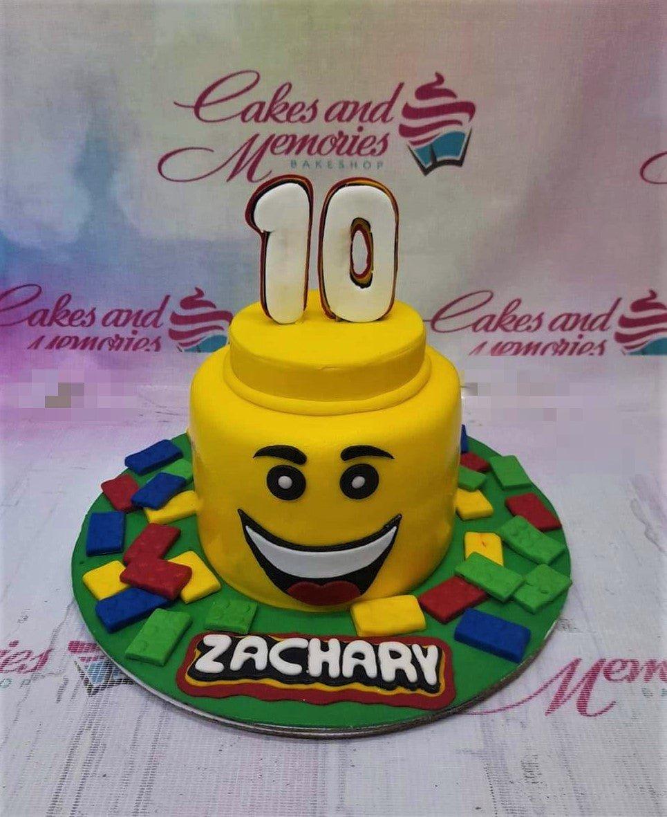 Yellow Lego head cake with a large number 10 topper and colorful Lego brick decorations on a green base.