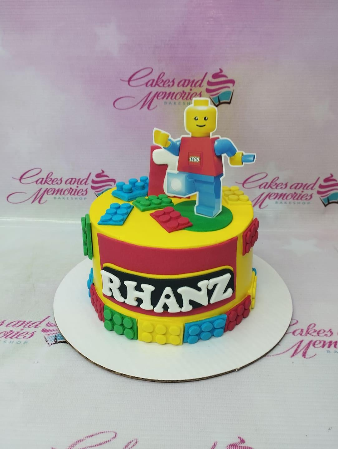 Yellow Lego themed birthday cake with a large Lego man printout topper and colorful fondant brick decorations.