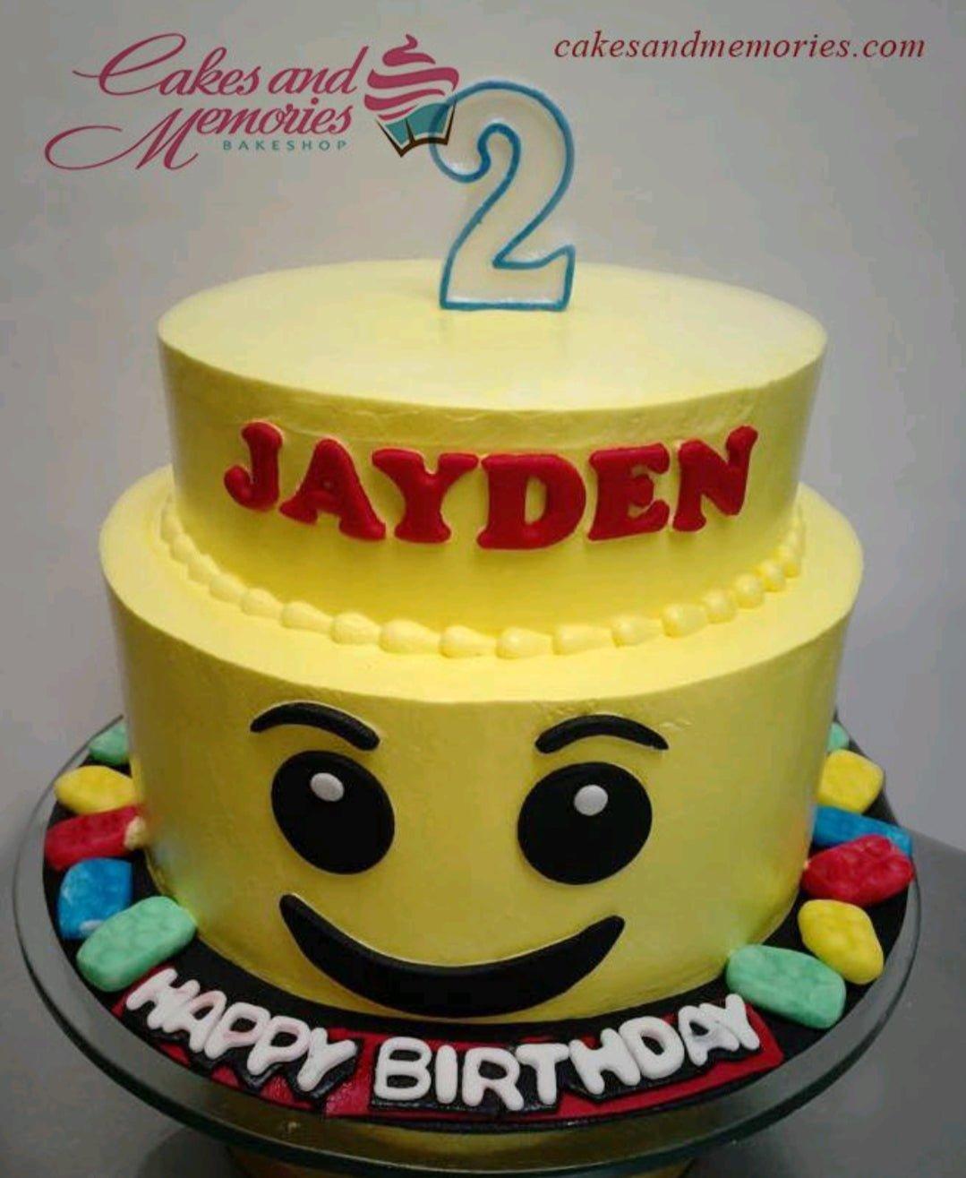 Yellow 2-tier Lego-themed birthday cake with a smiling Lego face on the bottom tier and red gumpaste letters for the name Jayden.