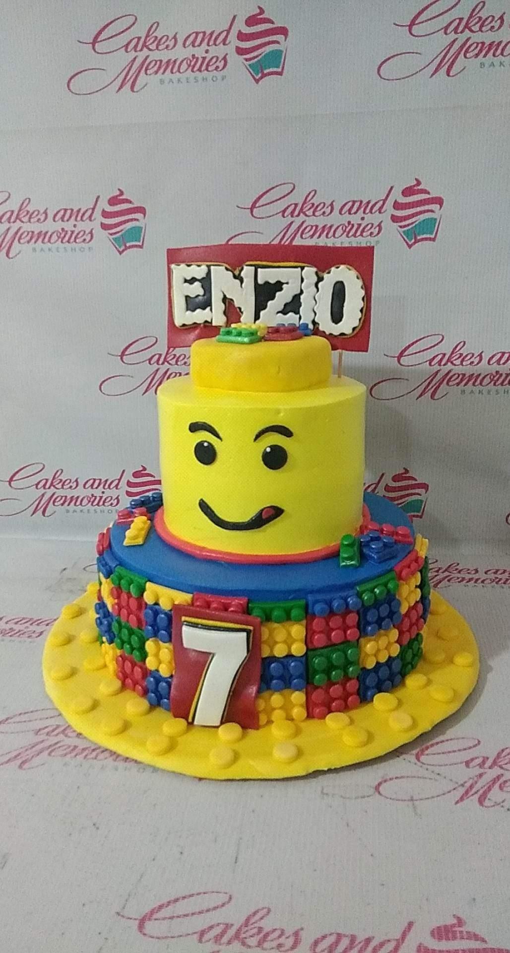 Two-tier Lego themed birthday cake with a yellow Lego head top tier and a blue bottom tier decorated with colorful Lego brick patterns.
