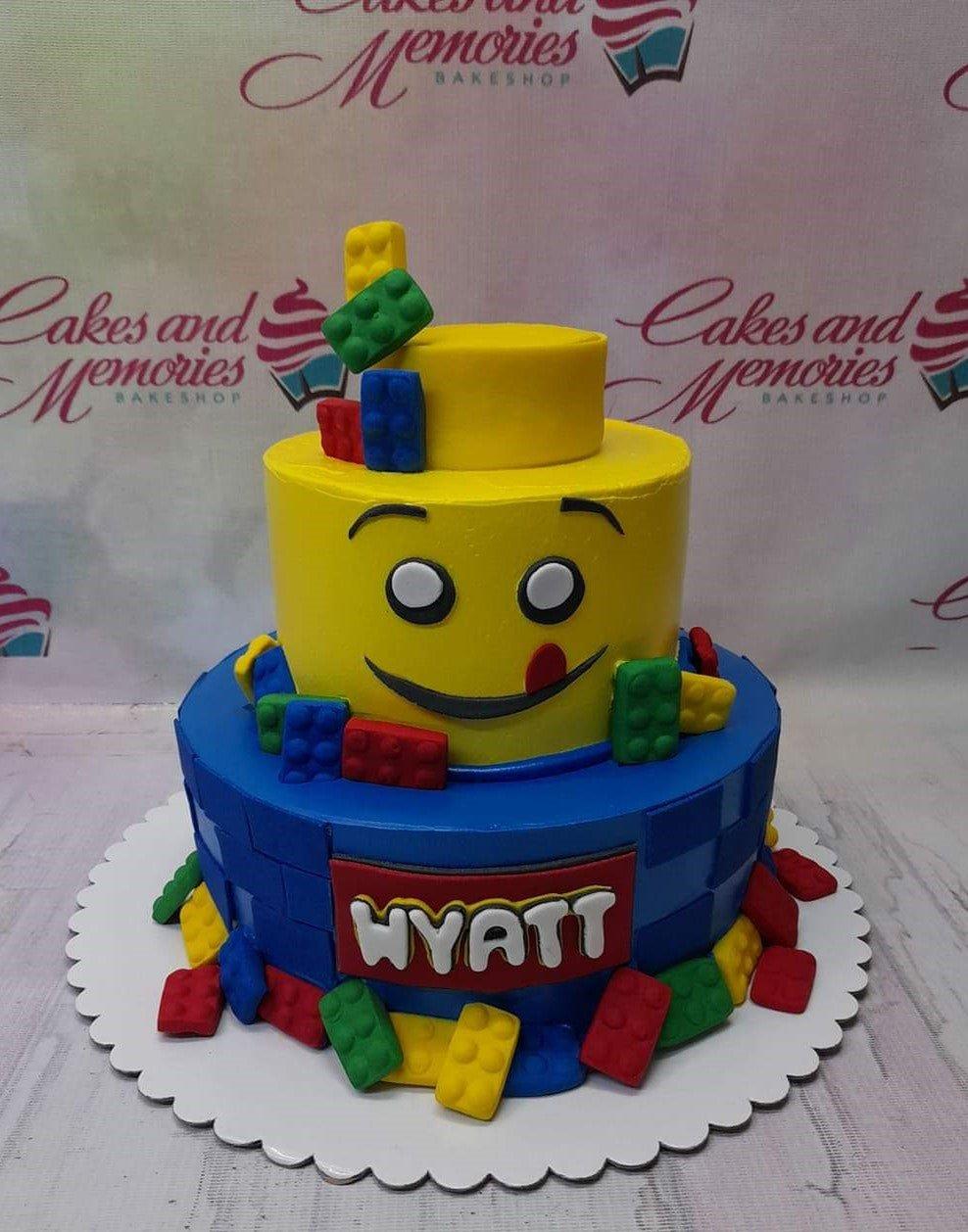 Two-tier Lego-themed birthday cake with a yellow Lego head top tier and blue brick-patterned bottom tier featuring colorful fondant blocks.
