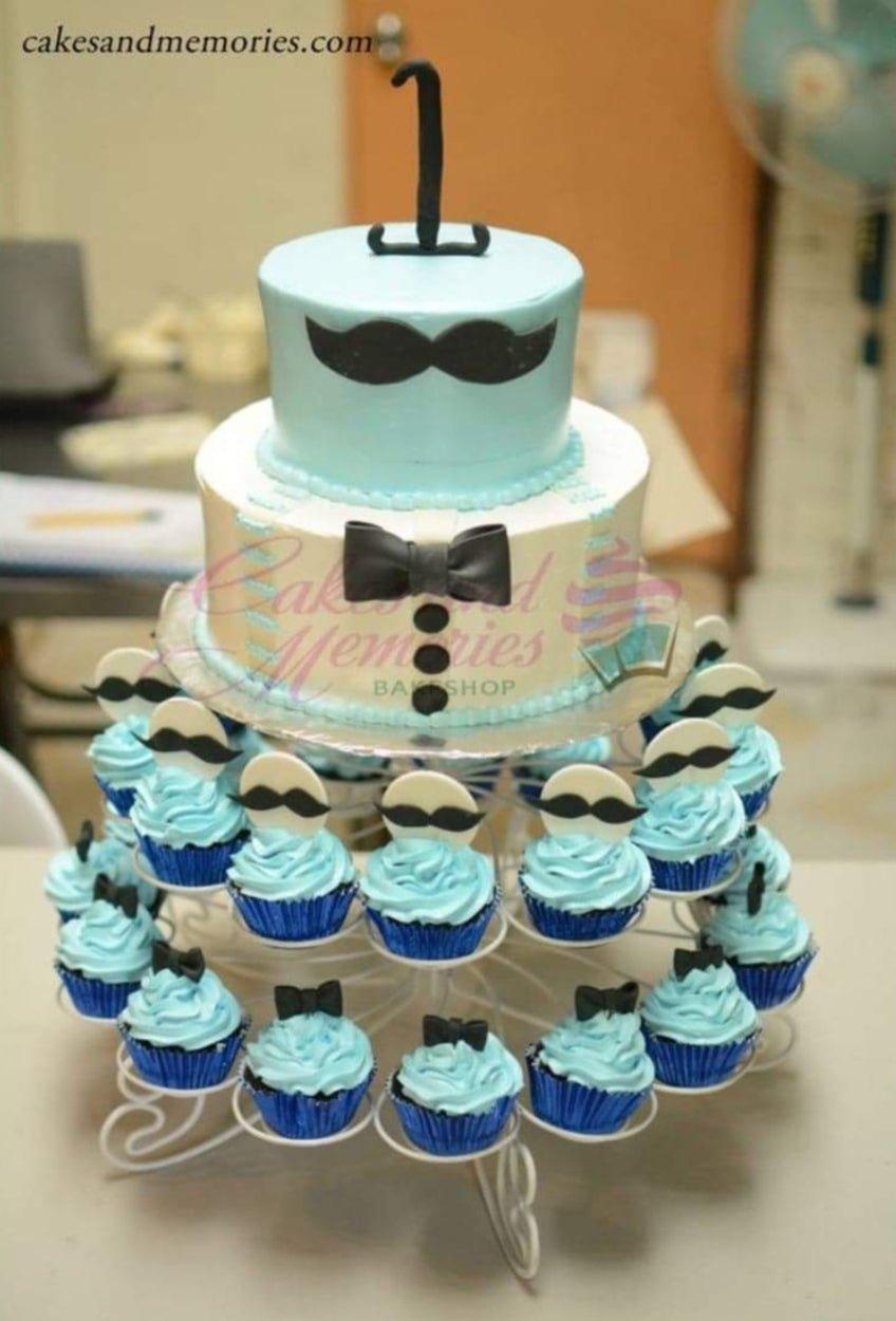 Two-tier light blue and white tuxedo cake with black mustache and bow tie toppers, surrounded by matching cupcakes.