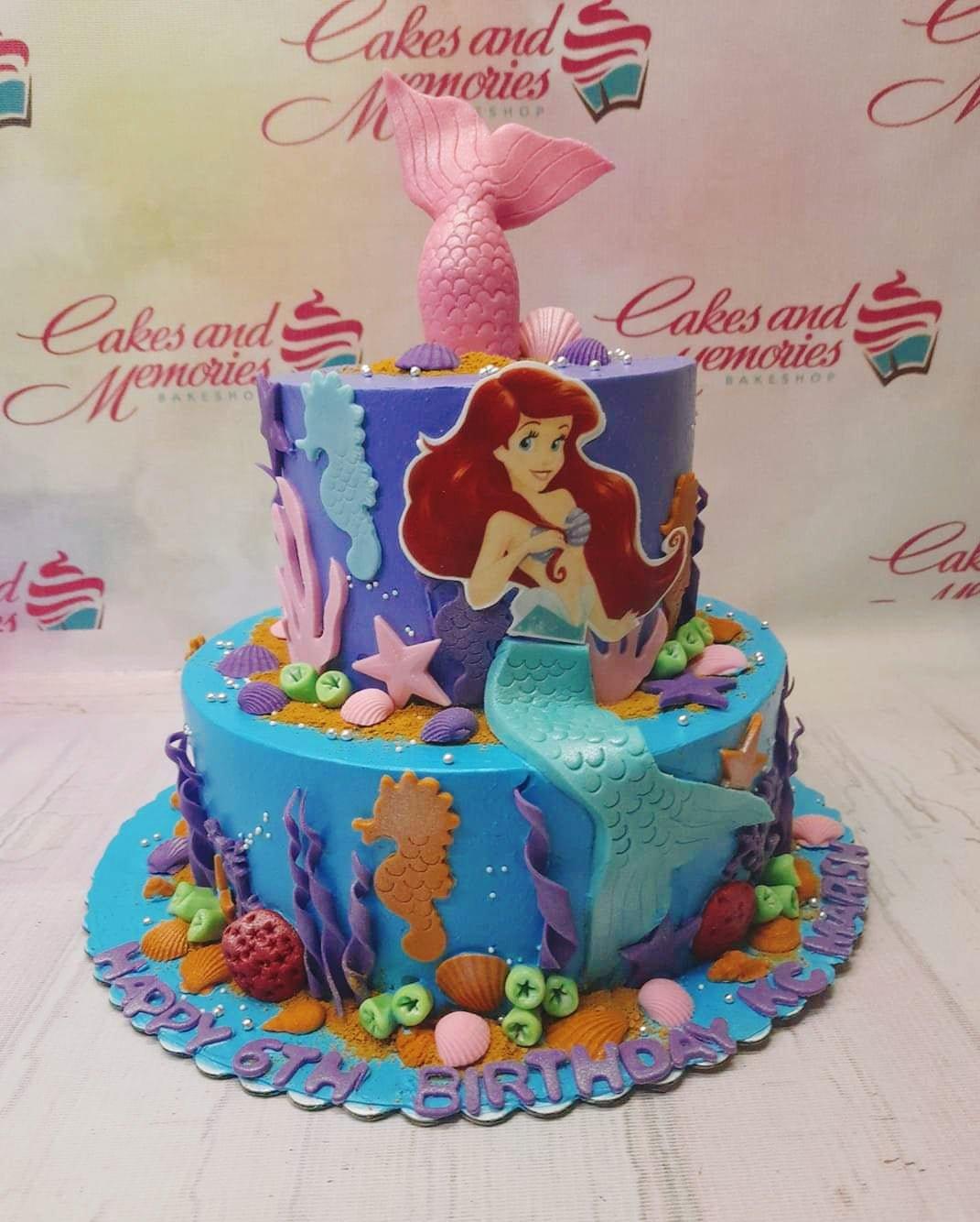 Two-tier Little Mermaid themed cake in purple and blue with Ariel printout, fondant mermaid tail, and sea life decorations.