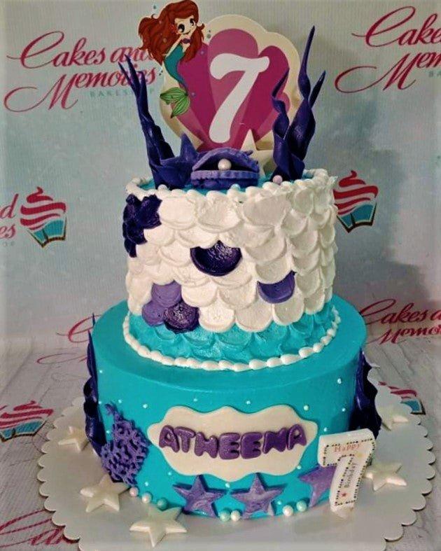 Two-tier Little Mermaid themed cake with white scale icing, teal base, and Ariel printout toppers.