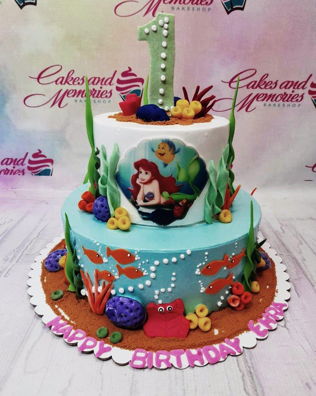 Two-tier Little Mermaid themed cake with Ariel printout, fondant sea creatures, and edible sand on a white base board.