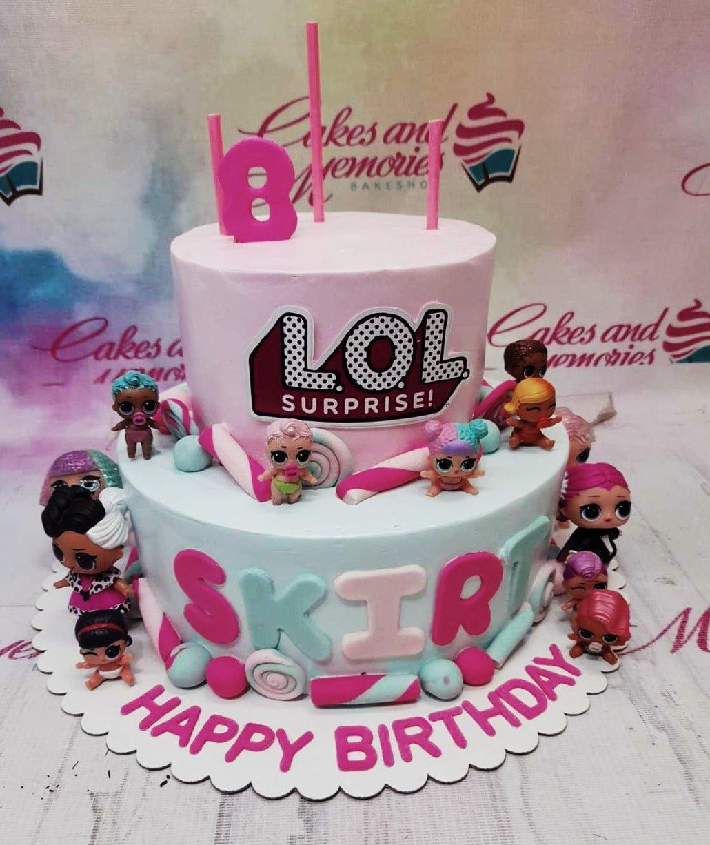 Two-tier LOL Surprise themed birthday cake in pink and light blue with various character figurines and candy-shaped fondant decorations.